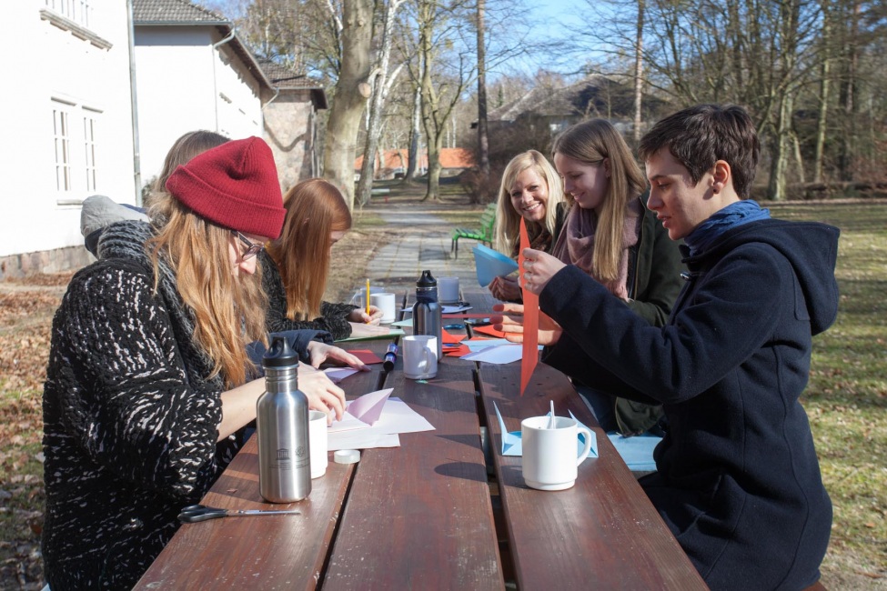 Sechs kulturweit-Freiwillige sitzen an einem Tisch im Freien und falten Briefe.