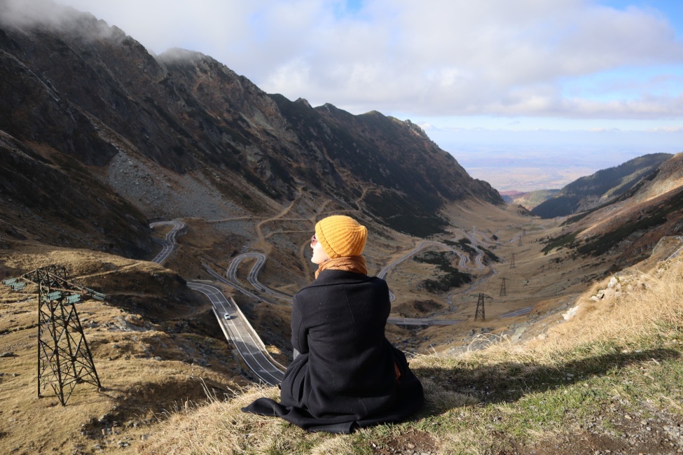 Freiwillige Angelique sitzt mit Mantel und Mütze in einer hügeligen Landschaft