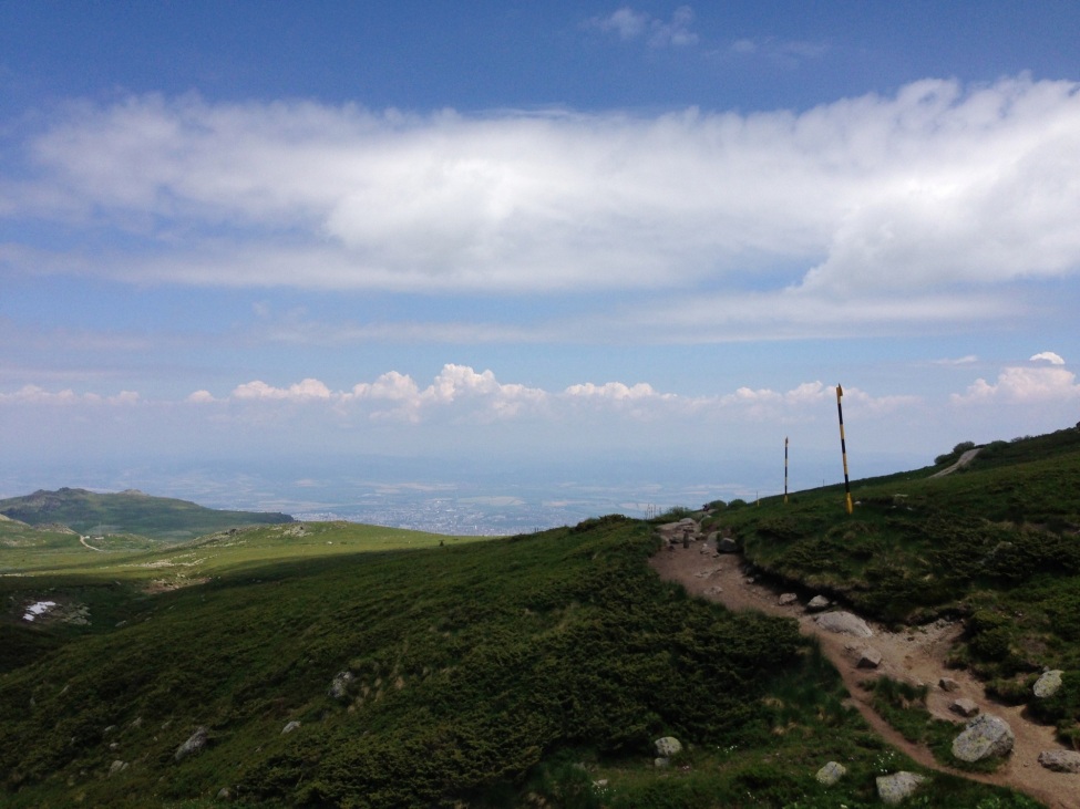 Blick über bulgarische Landschaft. Im Vordergund grüne Hügel, ein Weg schlängelt sich hindurch. Im Hintergrund im Tal eine Stadt